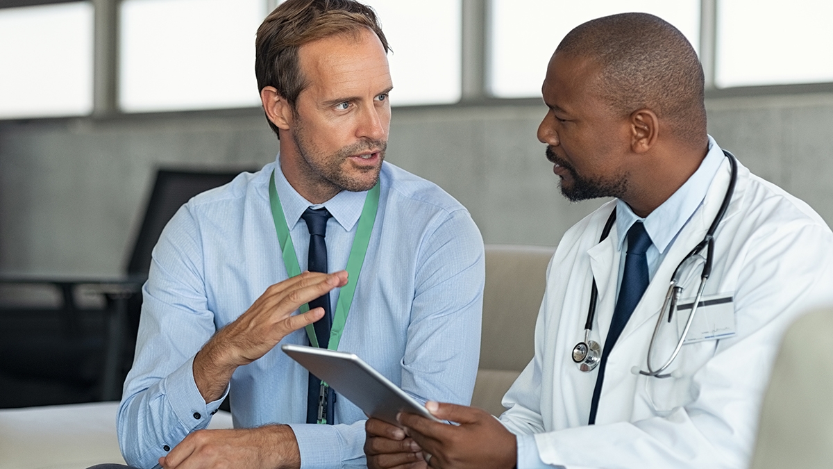 Pharmaceutical representative talking with doctor sitting on couch. African mature practitioner discussing results of the analysis with specialist while consulting diagnosis on digital tablet. Doctor in conversation with medical advisor in hospital room discussing patients report case.